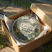 A decorative ornament in a box, featuring a scenic landscape with mountains, trees, and a waterfall.