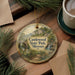 A decorative ornament featuring a scenic landscape of Castlewood State Park, Missouri, with a bridge and waterfalls. The ornament is placed on a wooden surface, surrounded by pine branches and a cup of coffee.