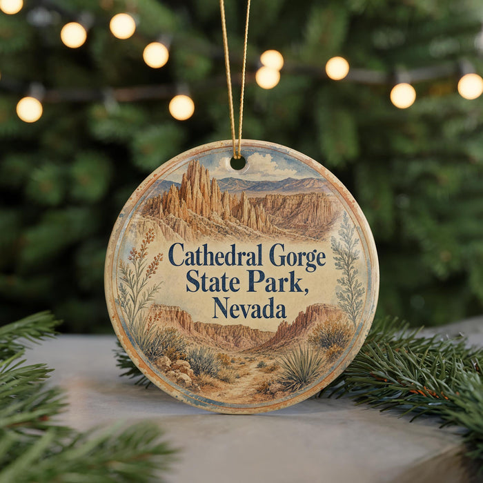 A round ornament with a scenic illustration of the Cathedral Gorge State Park in Nevada, featuring mountains, desert, and greenery, is displayed on a table with a blurred background of Christmas lights.