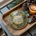 A decorative ornament featuring a scenic image of Cathedral State Park in West Virginia, with pine cones and a lit candle on a wooden tray.