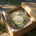 A decorative ornament in a box, featuring a scenic landscape with a waterfall and a bridge, with the text "Cathedral State Park, West Virginia" printed on it.