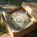 A decorative ornament in a box, featuring a scenic landscape with mountains, water, and greenery.