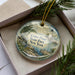 A decorative ornament in a box, featuring a scenic landscape with a lighthouse and a body of water.