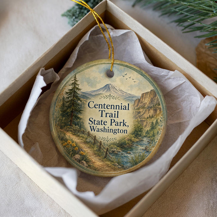 A round ornament with a scenic mountain landscape and trail, placed in a box with a white tissue paper lining.