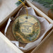A round ornament with a scenic mountain landscape and trail, placed in a box with a white tissue paper lining.