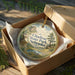 A round ornament in a box, featuring a scenic landscape of Chadron State Park, Nebraska.