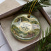 A decorative ornament in a box, featuring a scenic landscape with a waterfall and a bridge.