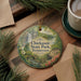 A decorative ornament featuring a scenic landscape with a waterfall, surrounded by pine branches and flowers, placed on a wooden surface next to a cup of coffee.