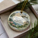 A decorative ornament with a scenic landscape of Chimney Rock State Park in North Carolina, featuring a mountain, pine trees, and a river.