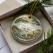 A decorative ornament with a scenic landscape featuring a deer, pine trees, and a body of water, placed in a box with a pine branch.
