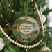 A round ornament hanging on a Christmas tree, featuring a scenic landscape with a river, mountains, and a deer.