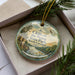 A decorative ornament with a scenic landscape of cliffs, trees, and a river, placed inside a box with a pine branch.