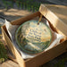 A round ornament in a box, featuring a scenic landscape with trees, a river, and a bird in flight.
