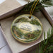 A decorative ornament in a box, featuring a scenic landscape with a bird and a house, surrounded by greenery.