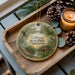 A round ornament with a painting of a park scene hangs on a wooden tray, accompanied by pine cones and a lit candle.