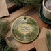 A round ornament with a scenic landscape of Colleton State Park, South Carolina, featuring a bird and a bridge, is displayed on a wooden surface alongside a cup of coffee and some pine branches.