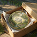 A decorative ornament in a brown box, featuring a scenic landscape with a bird and trees, with the text "Colleton State Park, South Carolina" printed on it.