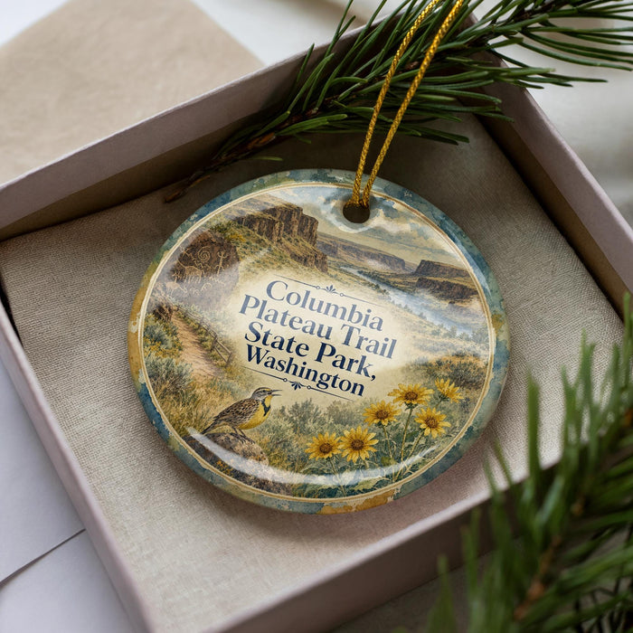 A decorative ornament in a box, featuring a scenic landscape with mountains, a bird, and flowers.