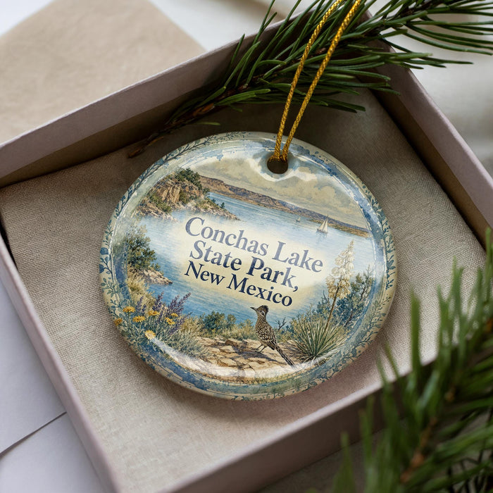 A decorative ornament in a box, featuring a scenic landscape with a lake, mountains, and a bird, along with the text "Conchas Lake State Park, New Mexico".