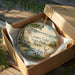 A decorative ornament in a box, featuring a scenic landscape with a lake, trees, and wildlife, including a deer and a bird.