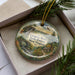 A decorative ornament in a box, featuring a scenic landscape with a bridge, trees, and a bird in flight.
