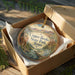 A decorative ornament in a box, featuring a scenic landscape with mountains, trees, and a river.