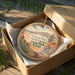 A round ornament in a box, featuring a scenic landscape with mountains, trees, and a desert-like environment.