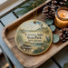 A decorative ornament with a lighthouse and beach scene, placed on a wooden tray next to pine cones and a lit candle.