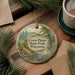 A decorative ornament with a scenic landscape of Cross Plains State Park, Wisconsin, featuring a winding path, pine trees, and wildflowers. The ornament is hanging on a wooden surface, surrounded by pine branches and a cup of coffee.