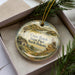 A decorative ornament in a box, featuring a landscape with a windmill, mountains, and a bird, with the text "Cross Ranch State Park, North Dakota" printed on it.