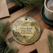 A decorative ornament featuring a windmill, a bird, and a rural landscape, with the text "Cross Ranch State Park, North Dakota" printed on it. The ornament is placed on a wooden surface, surrounded by pine branches and a cup of coffee.