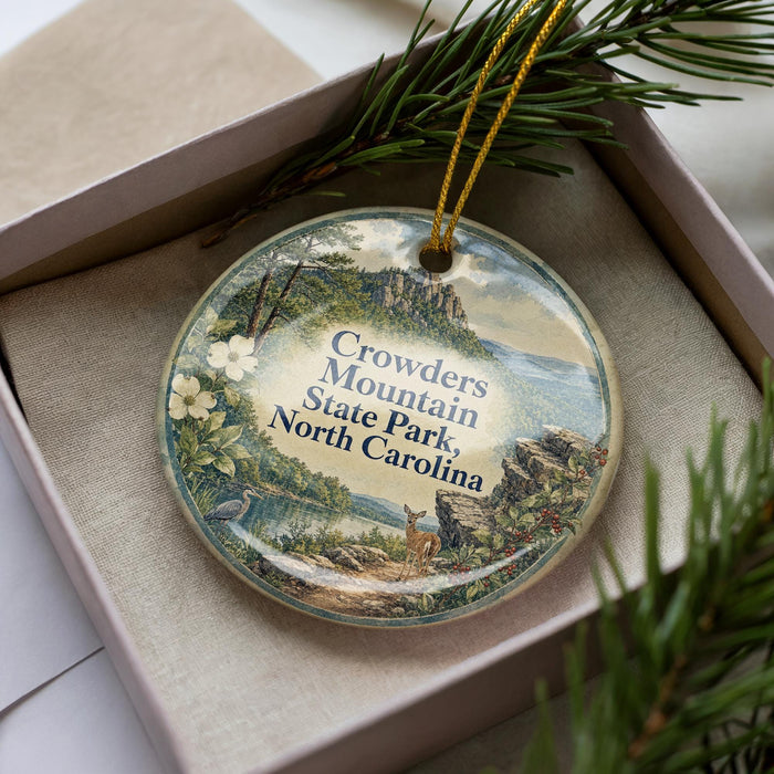 A decorative ornament in a box, featuring a scenic landscape with mountains, trees, and wildlife.