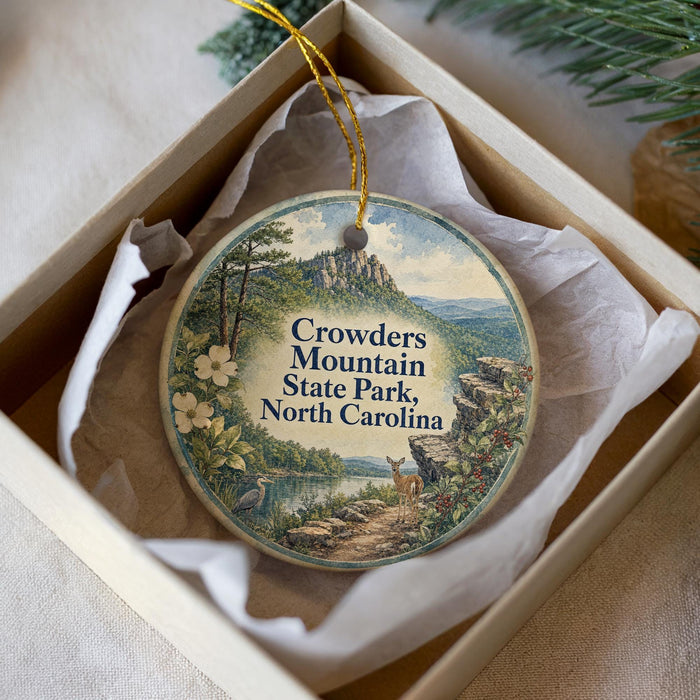 A decorative ornament in a white box, featuring a scenic landscape with mountains, trees, and a river.