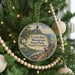 A round ornament hanging on a Christmas tree, featuring a scenic landscape with mountains, trees, and a winding path.