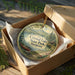 A round ornament in a wooden box, featuring a scenic landscape with a canoe and mountains, with the text "Crystal Lake State Park, Vermont" displayed on it.