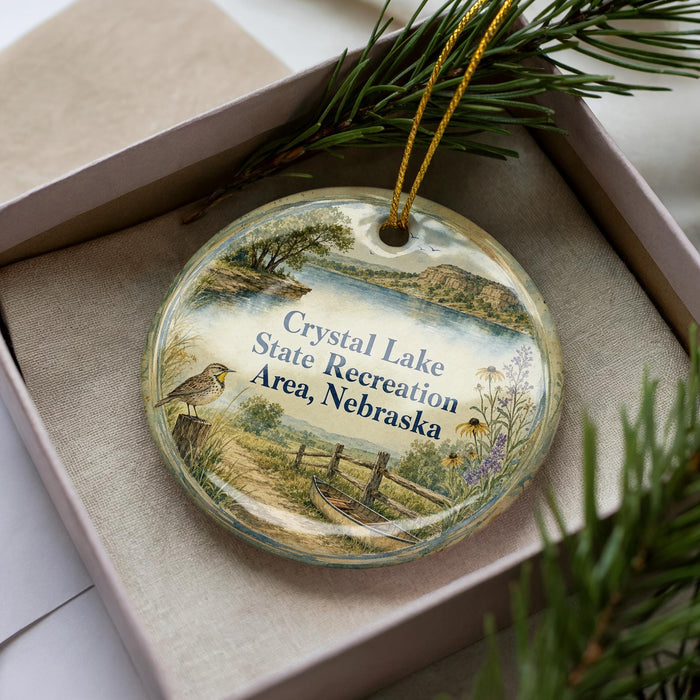 A decorative ornament in a box, featuring a scenic landscape with a lake, trees, and a bird.