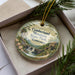 A decorative ornament with a scenic landscape of a waterfall and a house, placed inside a box with pine branches.