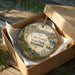 A decorative ornament in a box, featuring a scenic landscape with mountains, a river, and wildlife, including a bison and a duck.