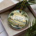 A decorative ornament in a box, featuring a scenic landscape with a body of water, trees, and mountains.