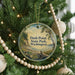 A circular ornament hanging on a Christmas tree, featuring a scenic landscape with a body of water and trees.