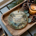 A decorative ornament featuring a scenic landscape with a bridge, lighthouse, and pine trees, placed on a wooden tray alongside pine cones and a lit candle.
