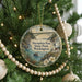 A round ornament hanging on a Christmas tree, featuring a scenic landscape with a bridge, lighthouse, and water.