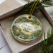 A decorative ornament in a box, featuring a scenic landscape with a waterfall and a bird, is surrounded by pine branches.