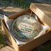 A wooden ornament in a box, featuring a scenic landscape with mountains, trees, and a river.