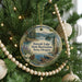 A round ornament hanging on a Christmas tree, featuring a scenic landscape with a lake, mountains, and pine trees.
