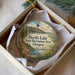 A decorative ornament in a box, featuring a scenic landscape with a lake, mountains, and trees.