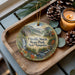 A decorative ornament featuring a scenic landscape of Dixville Notch State Park, New Hampshire, with pine cones and a lit candle on a wooden tray.