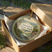 An ornament in a box, featuring a scenic landscape with mountains, trees, and a small cabin, with the text "Douthat State Park, Virginia" displayed on the ornament.