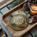 A decorative ornament featuring a scenic image of Droop Mountain Battlefield State Park, West Virginia, with a candle and pine cones on a wooden tray.