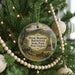 A round ornament hanging on a Christmas tree, featuring a scenic landscape with mountains, trees, and a cannon.